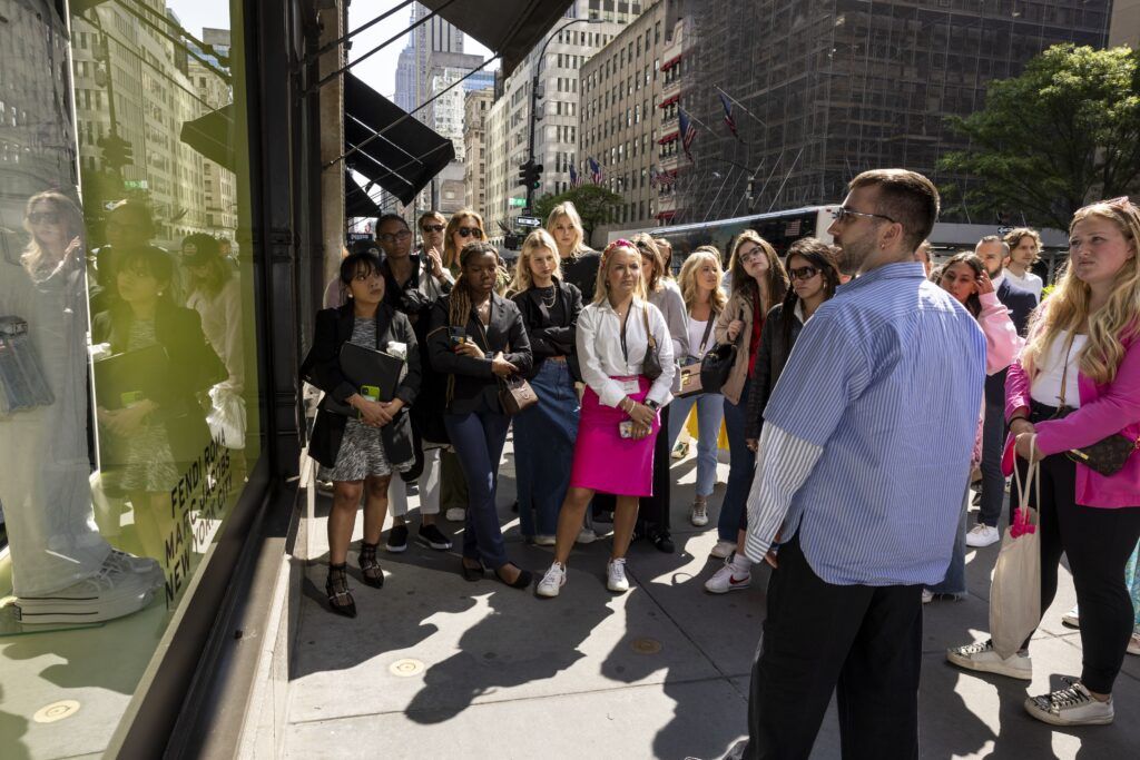 A group of college students listens intently to a man in a blue striped shirt while standing on a sidewalk outside a storefront.