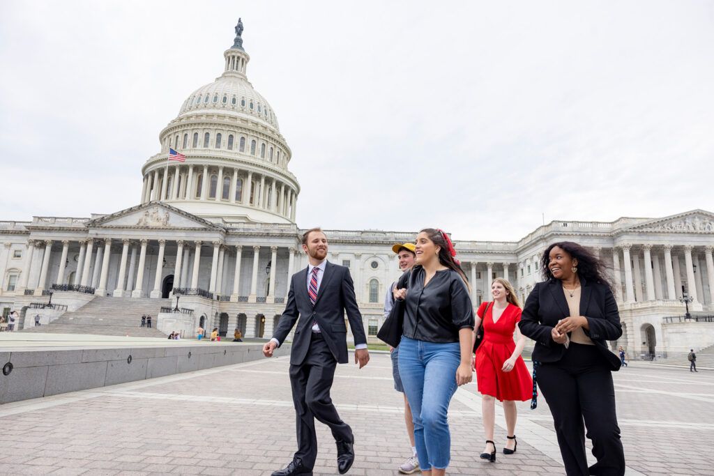 A group of formally dressed students walks past the Capitol Building in Washington D.C. The building is large, white, and feature classic architecture with a domed spire at the top.