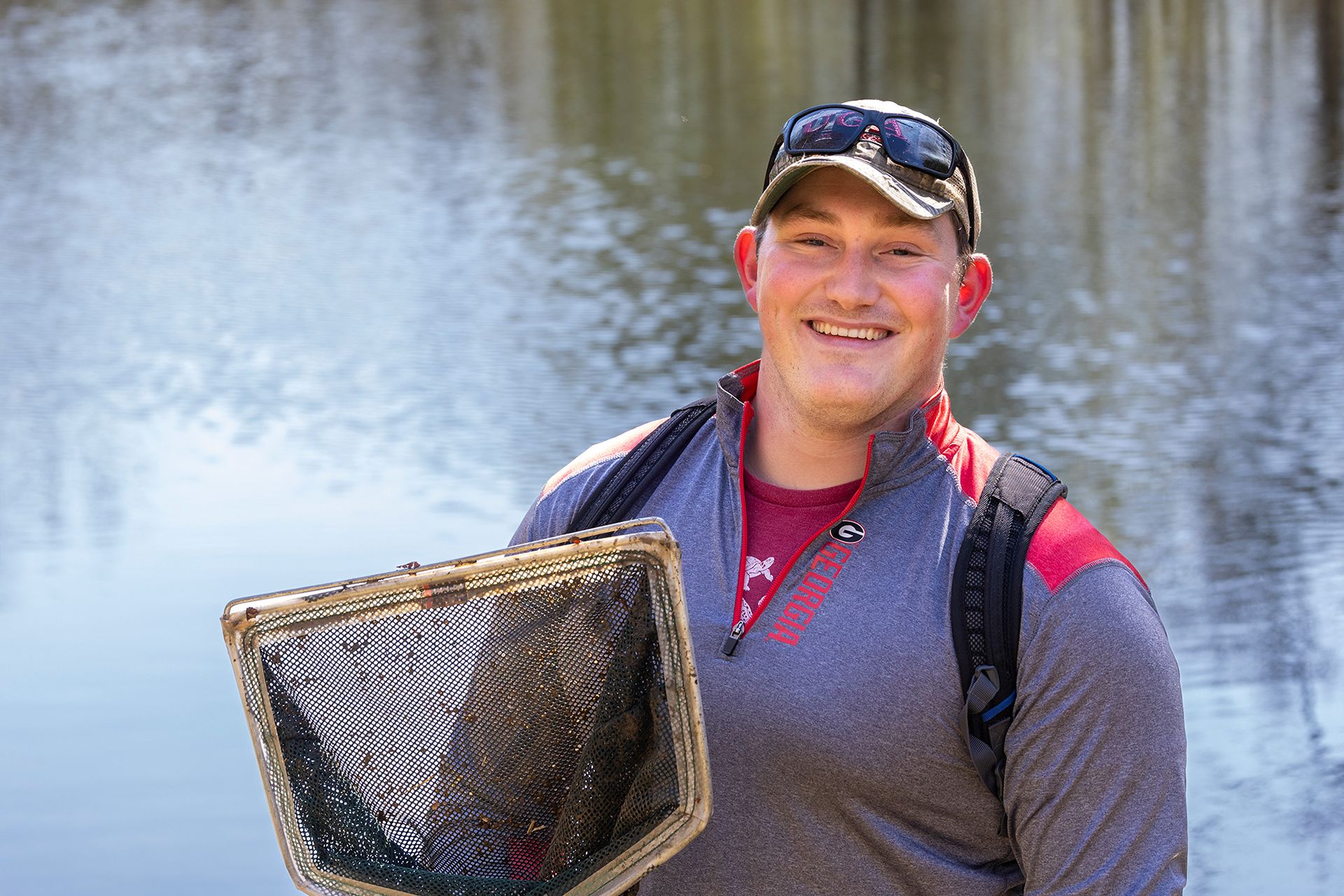 A closeup of a young man in a long-sleeved University of Georgia shirt and baseball cap outside in front of a lake. The man is holding a mesh net.