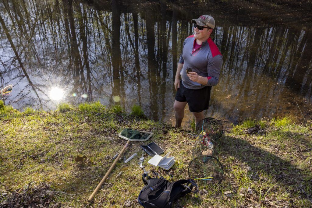 A man wearing dark shorts, a baseball cap, and a blue and red shirt stands on the grass by a river. He is holding a tiny turtle. Scattered on the ground are a backpack, a green mesh net attached to a pole, a notebook, and other small tools.