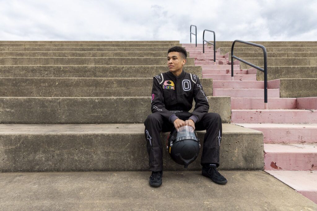 A young Black man sits on steps outdoors and looks off to the side. He is wearing a black racing jumpsuit and holds his black racing helmet in both hands between his knees.