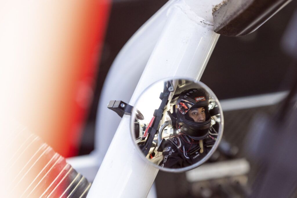 A man wearing a black racing helmet sits in his race car. His face is reflected in the rearview mirror.