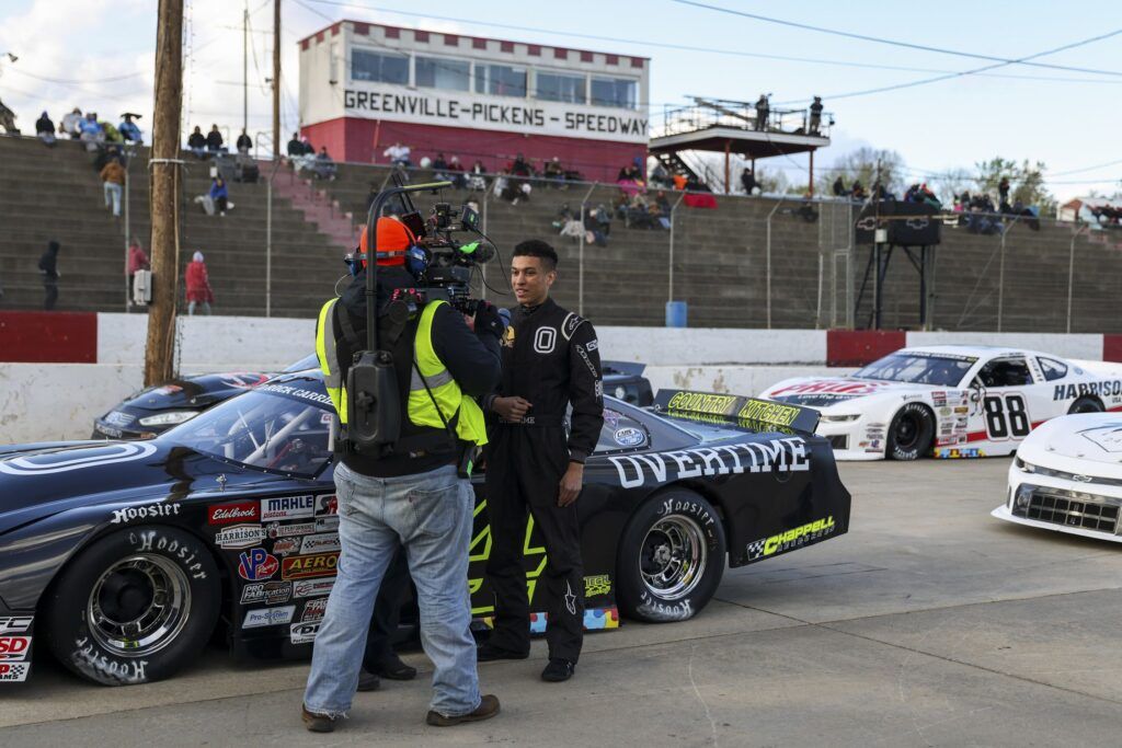 A young Black man in a black racing jumpsuit stands beside a black race car covered in stickers. He is being interviewed by a larger man holding a camera on his shoulder. Behind them are several fans seated in bleachers outdoors at the Greenville-Pickens Speedway.