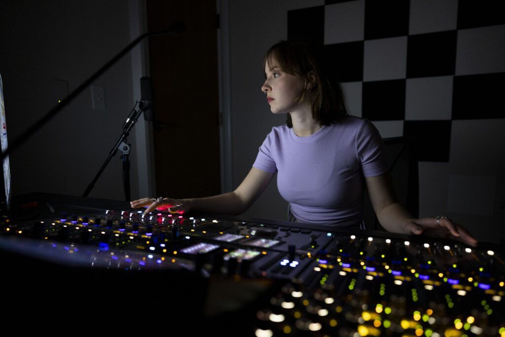 A woman in light purple shirt in a production studio's darkened music room operates the sound board.