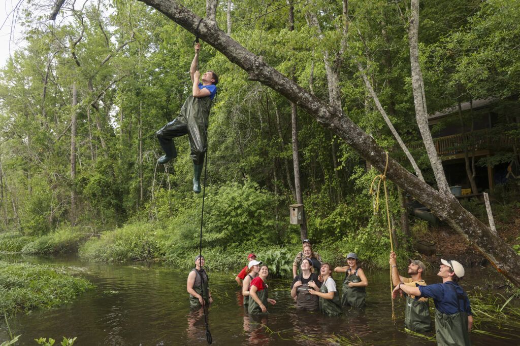 Several people in dark waders stand waist deep in a dark, swampy water. A man in dark green waders hoist himself up a rope that is tied to a thick tree limb above the water. There are many bright green trees in the background.