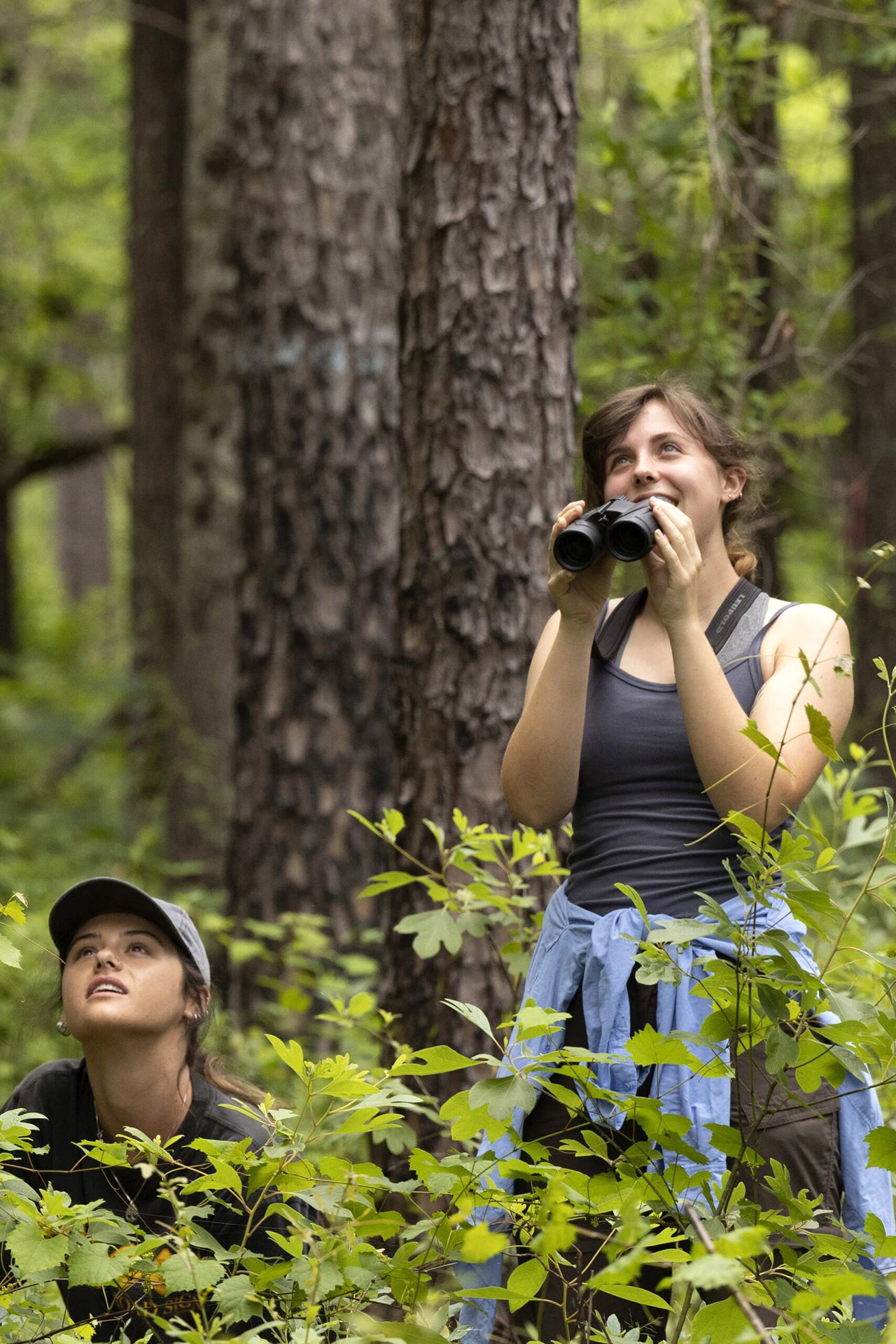 Two young women in the middle of the nature reserve known as the Savannah River Site look upwards towards the sky. They are wearing dark clothing and one is holding binoculars but not looking through them.