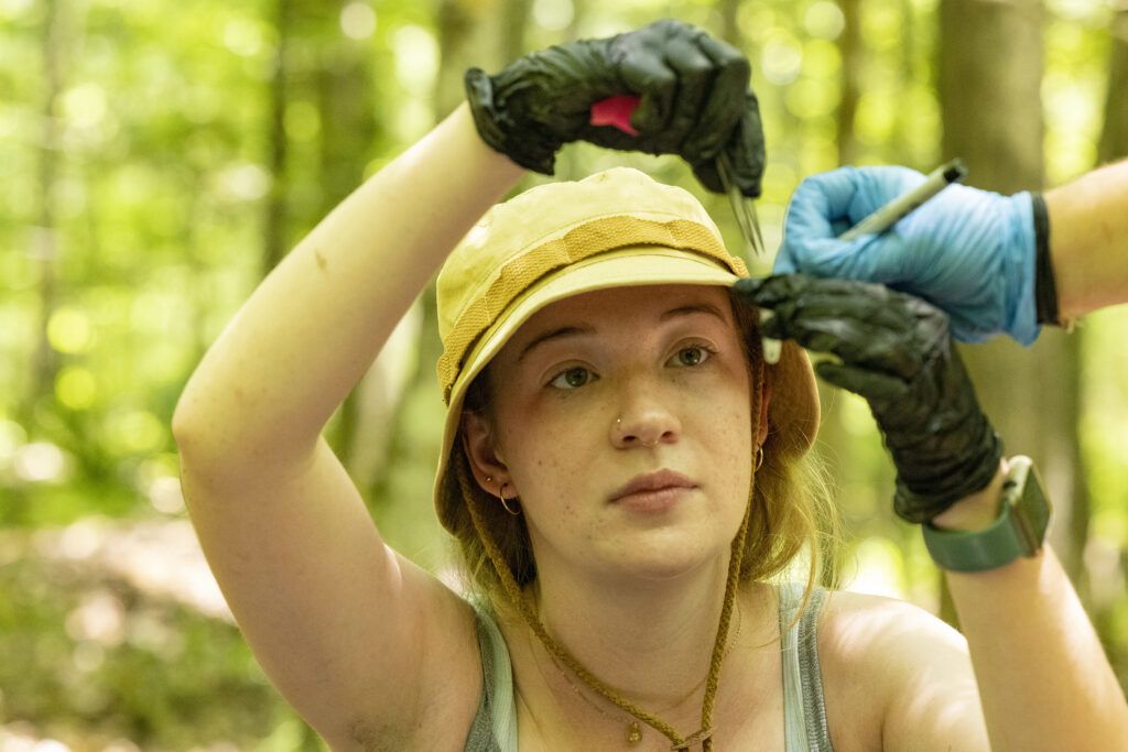 A closeup of a young woman in the middle of a forest studying a test tube. She is wearing black gloves and a yellow bucket hat.