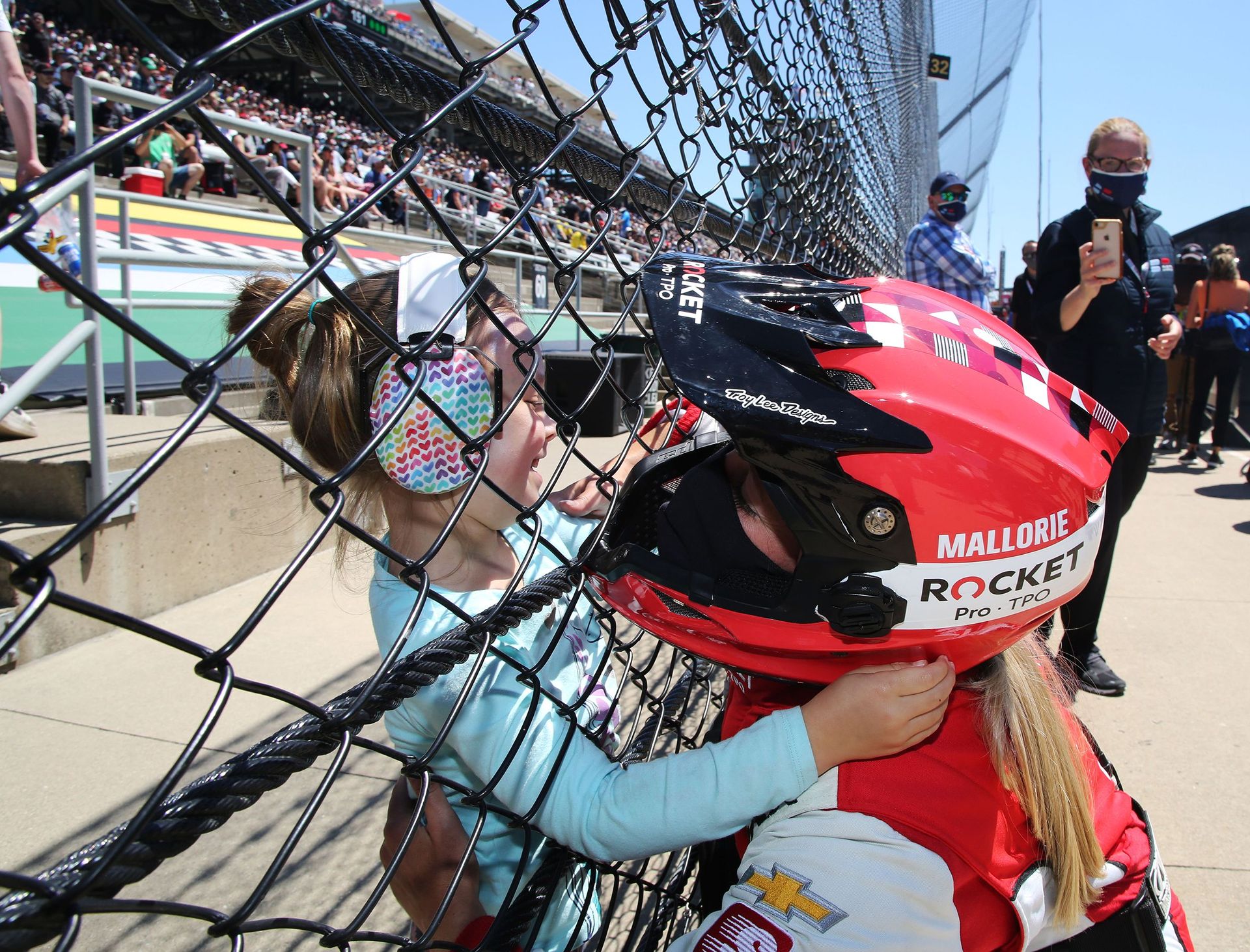 A woman wearing a red and black racing helmet with a blonde ponytail sticking out of the back puts her face close to a chain link fence that separates her from a little girl wearing headphones covered in hearts and a light blue shirt.