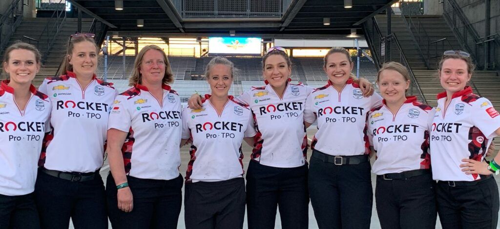 Eight young women stand side by side with their arms around each other posing for a group photo. They all wear the same uniform of white shirt that says Rocket Pro TPO and black pants.