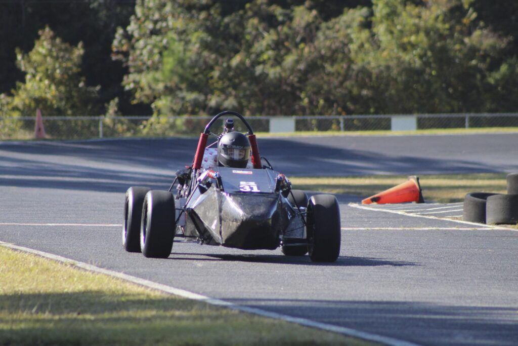 A man in a black racing helmet sits in a small mostly black Formula One vehicle as he drives around a track beside grass. An orange cone has fallen over in the background