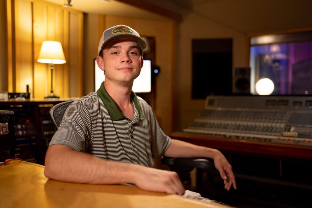 A close up of a young man in a baseball cap and striped shirt sitting in a music production studio.