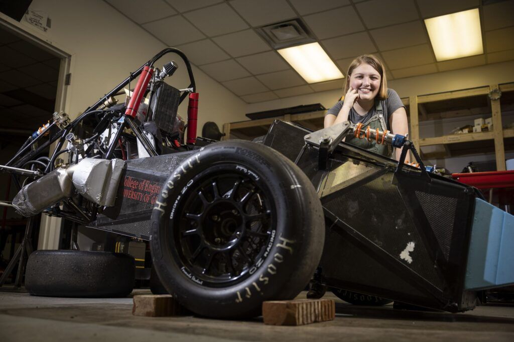 A smiling woman with bobbed blonde hair wears a grey T-shirt. She leans on the frame of a Formula One car that is under construction.