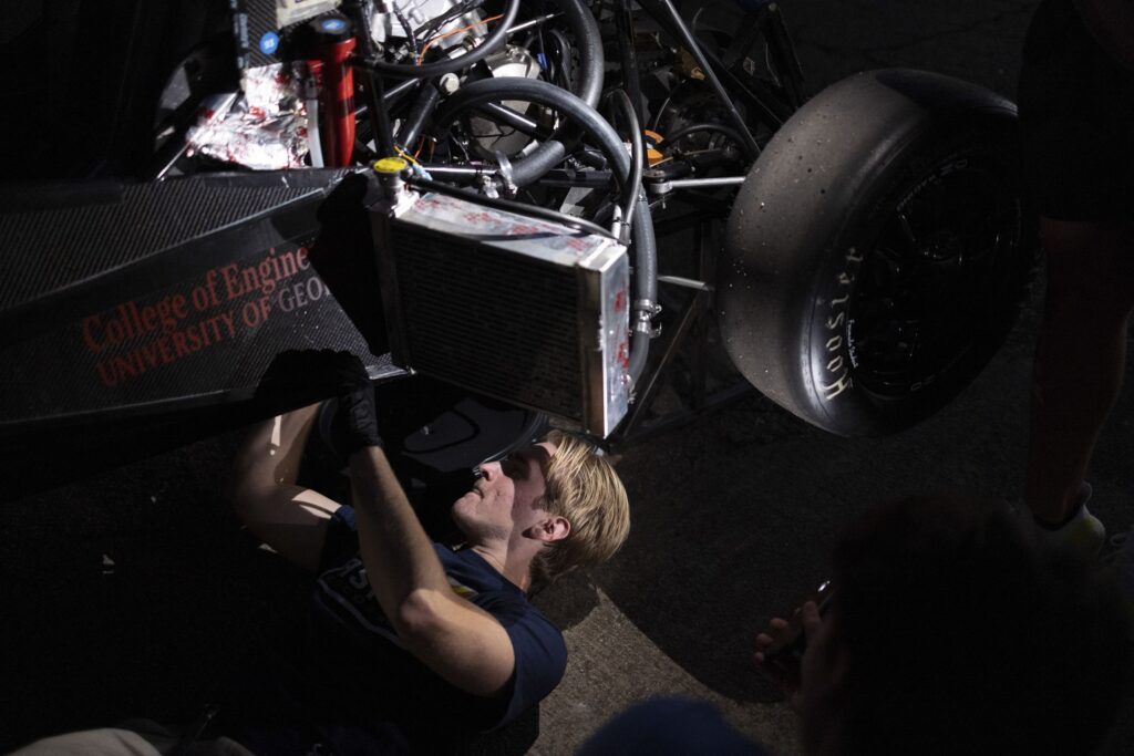 A blonde man in a black T-shirt is half visible from under a Formula One vehicle. His hands are raised, and he is working on the underside of the engine.