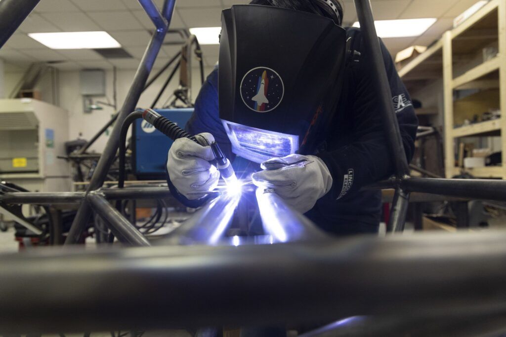 A man wearing a welding helmet and white gloves is bend over the frame of a vehicle welding a piece of metal