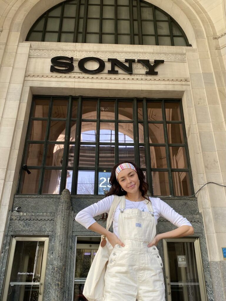 A woman in a white shirt and pale yellow overalls smiles while holding her hands on her hips. She is standing in front of a large arched doorway with the "Sony" logo near the top of the arch.