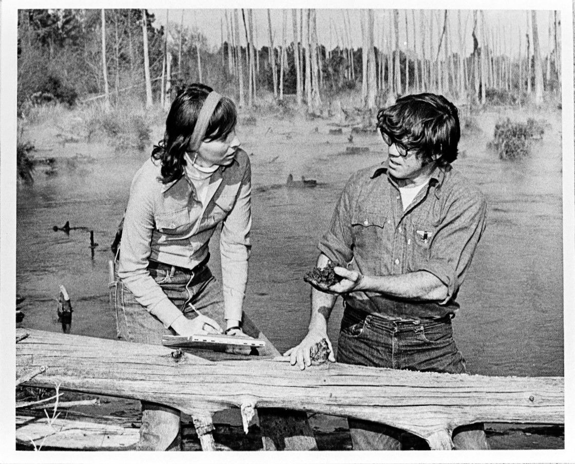 A black and white photo of a man and a woman on the side of a river. There is a large tree trunk laying on its side in front of them and many bare trees standing behind them.