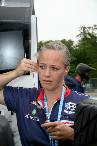 A blonde woman adjusts headphones in her right ear as she stands beside a van wearing a dark blue shirt with a red collar.