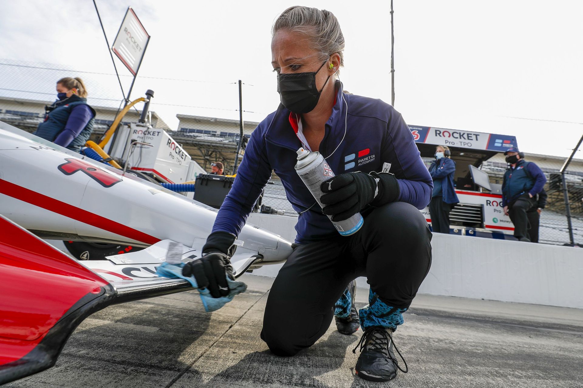 A blond woman wearing a black COVID-style mask, blue shirt and black pants knees beside the front of a red and white race car. She wipes the car with a blue cloth.