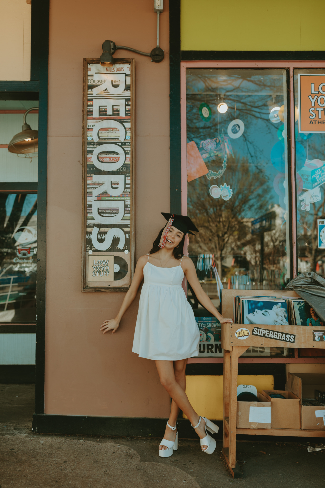 A young woman in a short white dress and black graduation cap poses in front a record store with her legs crossed and her arms against the store's wall.