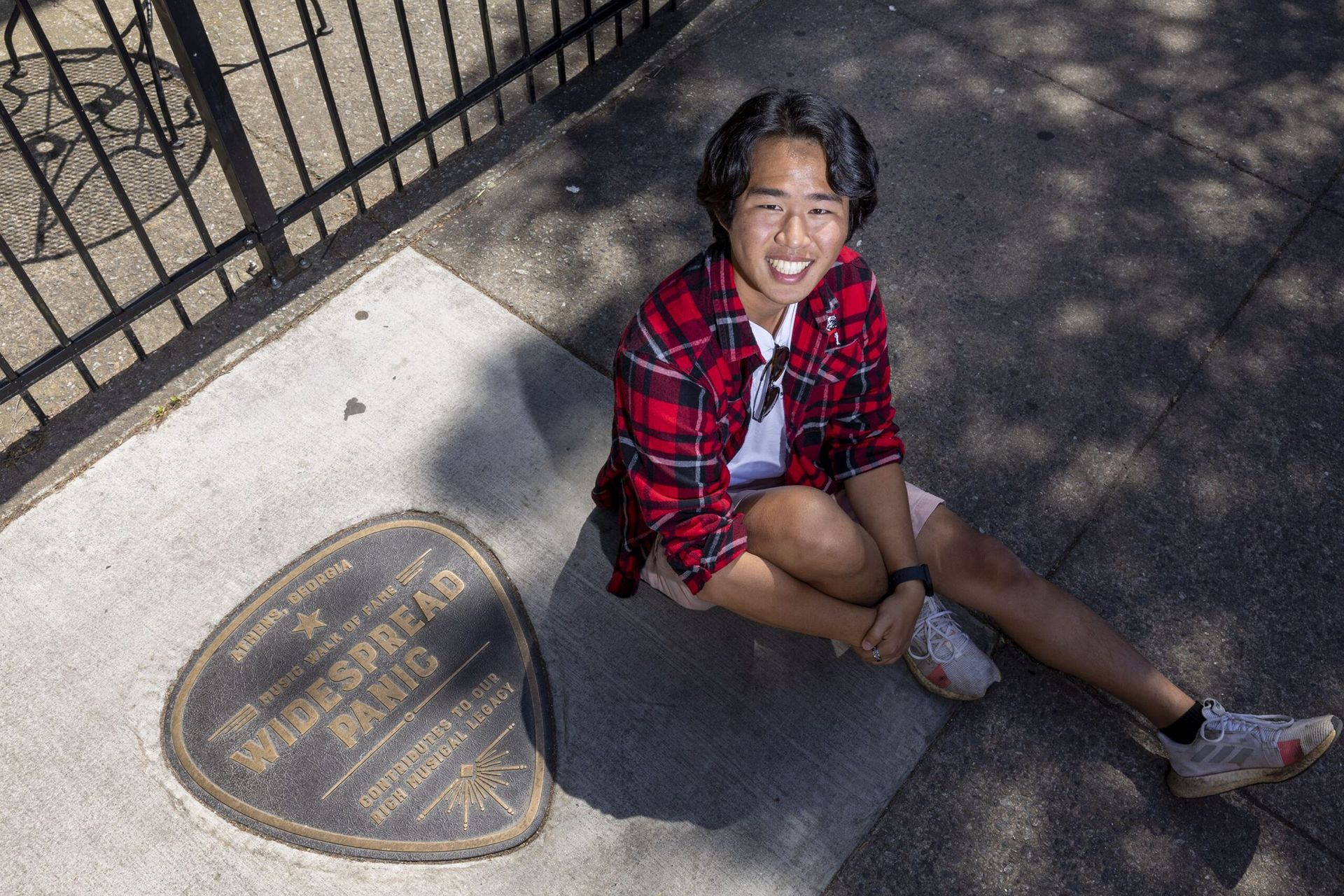 A young student sits on the pavement while smiling up at the camera. In the bottom left corner of the photo is guitar-pick shaped plaque dedicated to Widespread Panic.