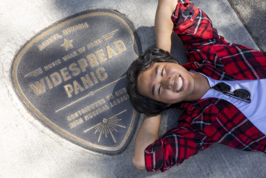 A smiling young man in a red and black plaid shirt lays on the ground with his hands behind his head. He is laying next to a guitar-pick shaped plaque dedicated to the band "Widespread Panic".