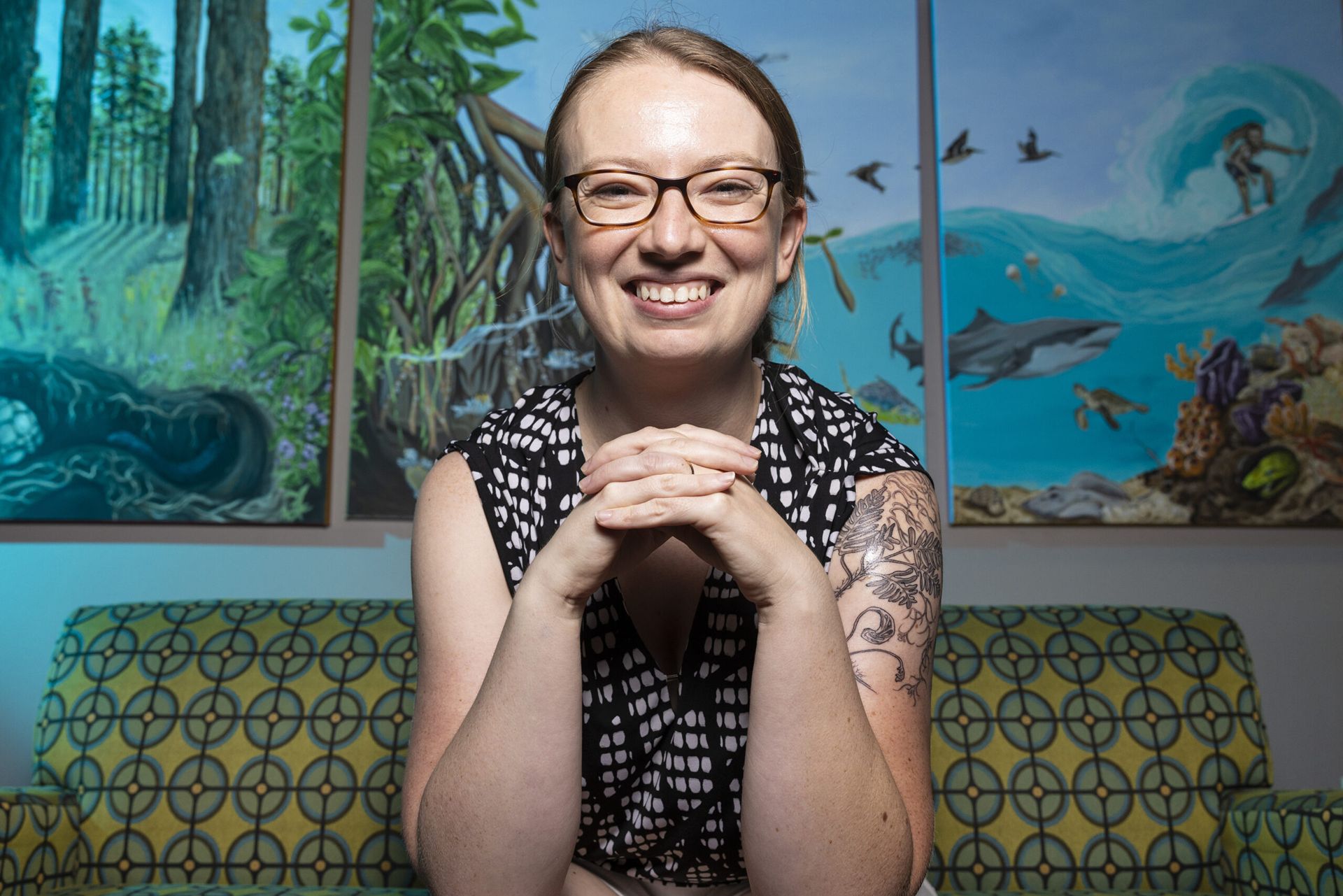 A close-up profile photo of Megan Marie Tomamichel. She is sitting on a short green and yellow couch and smiling at the camera. Her hands are clasped in front of her.