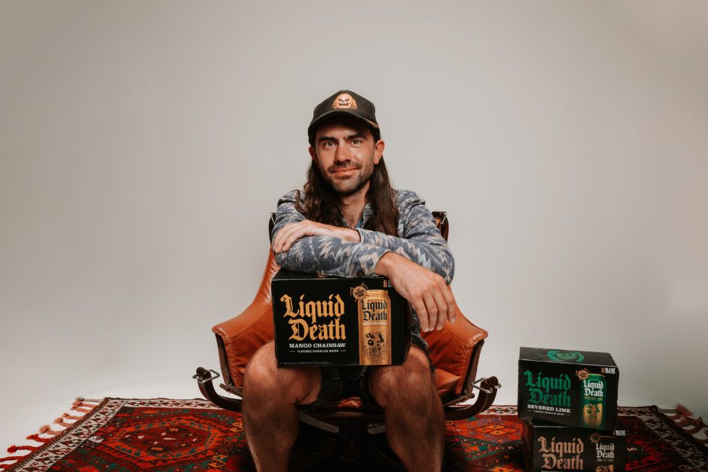 Andy Pearson wears a dark patterned shirt and baseball cap and sits in a modern orange chair. In his lap is a case of water with the label "Liquid Death Mango Chainsaw."