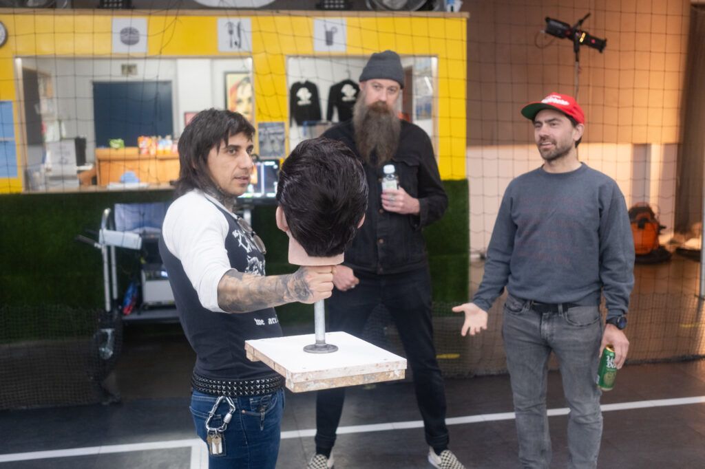 A group of people shoot a commercial for the company Liquid Death. A man in a black and white shirt holds a fake head mounted on a short steel pole. Two men in dark clothing look at him as they talk.