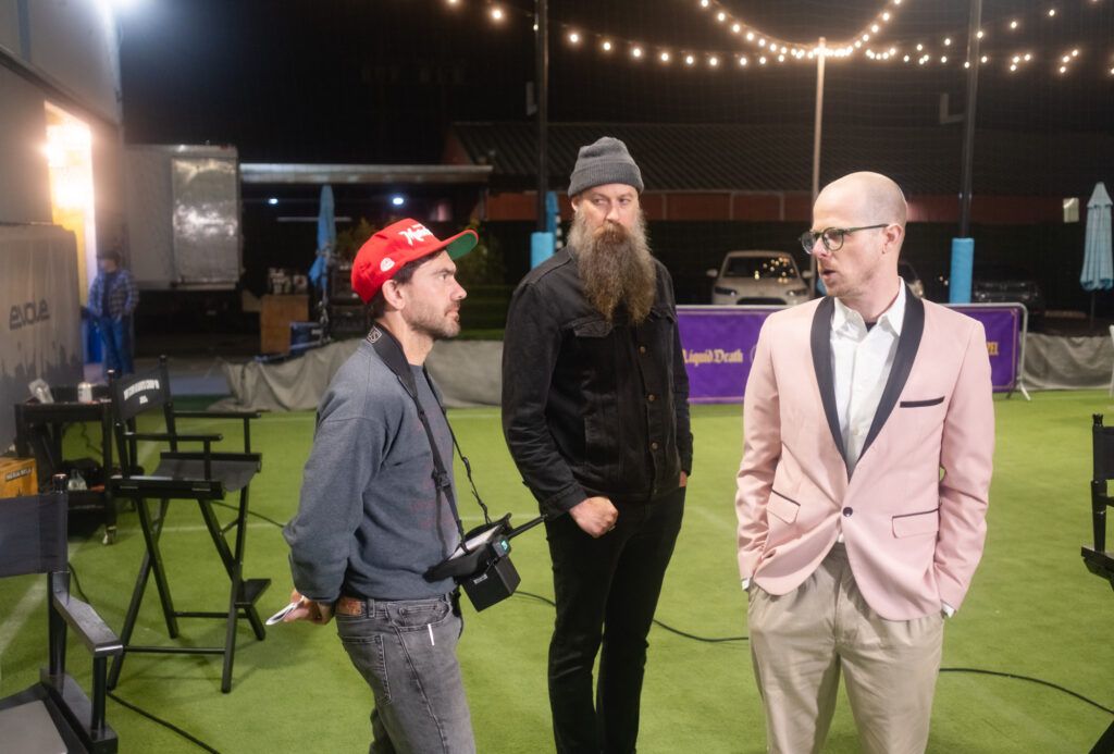 A group of people shoot a commercial for the company Liquid Death. One man stands with a boxy camera on a strap around his neck. The other two men look at him during their conversation.