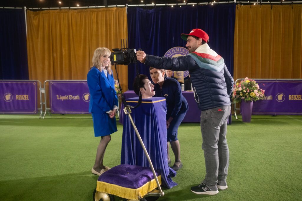 A group of people shoot a commercial for the company Liquid Death. One man is mostly hidden in a tall box covered with purple cloth. Only his head pokes out. Another man holds a camera above the first man's head. The last two people pose and make silly faces.