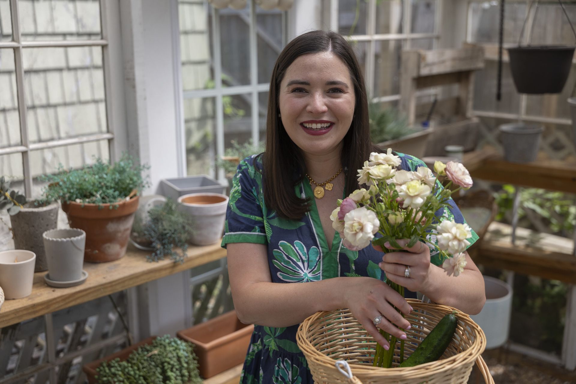 Ivy Odom smiles as she holds a small bouquet of flowers and a basket. She is standing in a greenhouse and is surrounded by many potted plants.