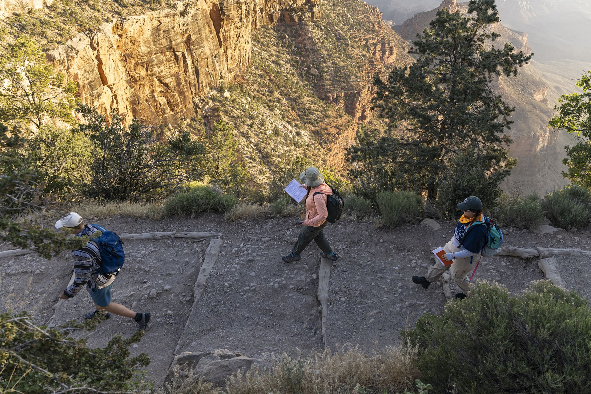 A high angle photo shows three people hiking on a mountain pathway with part of the Grand Canyon in the background.