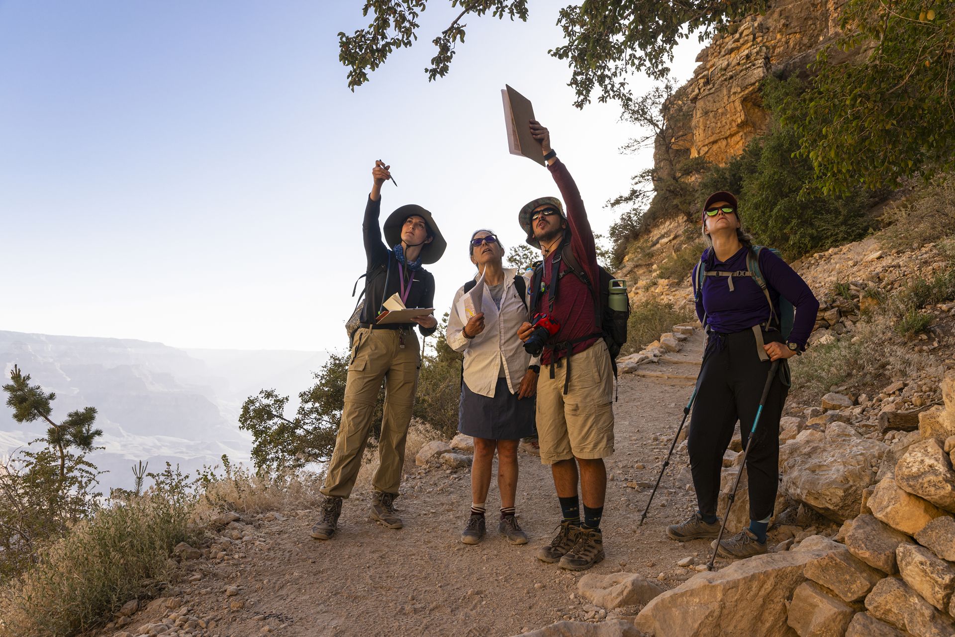 Four individuals in hiking clothes look up on a rocky trail with a hazy Grand Canyon in the background.