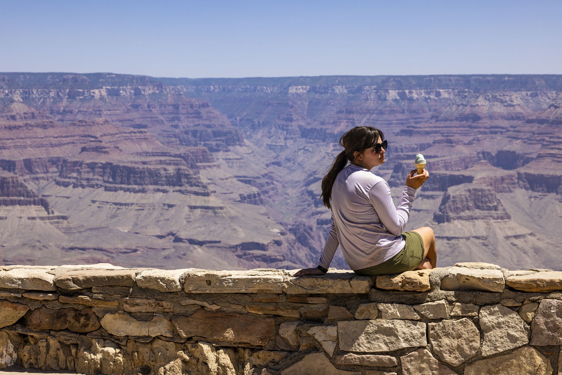 With the Grand Canyon in the background, a young women in sunglasses enjoys on ice cream cone.