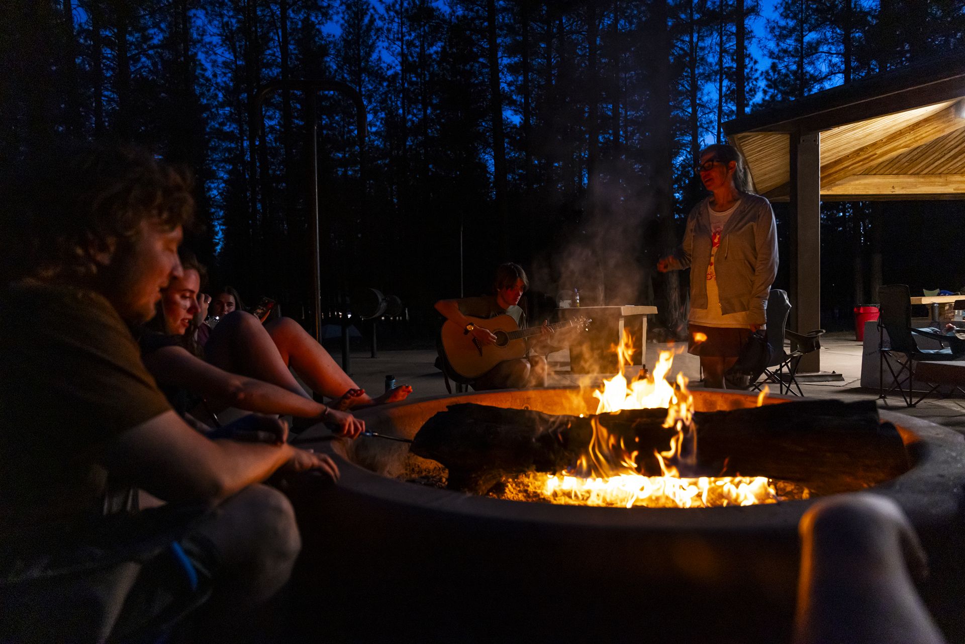 A log burns orange in a fire pit as barely lit faces stand into it. In the background someone is strumming a guitar.