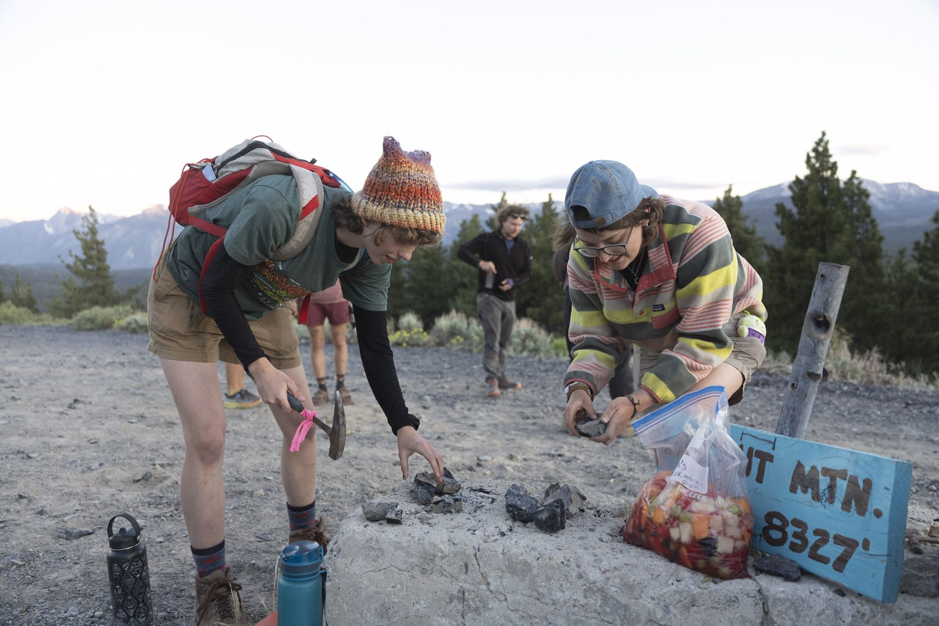 Two female students at the top of a mountain handle black rocks and a chisel at dusk. Near them is a 2 gallon Ziplock of fruit salad.