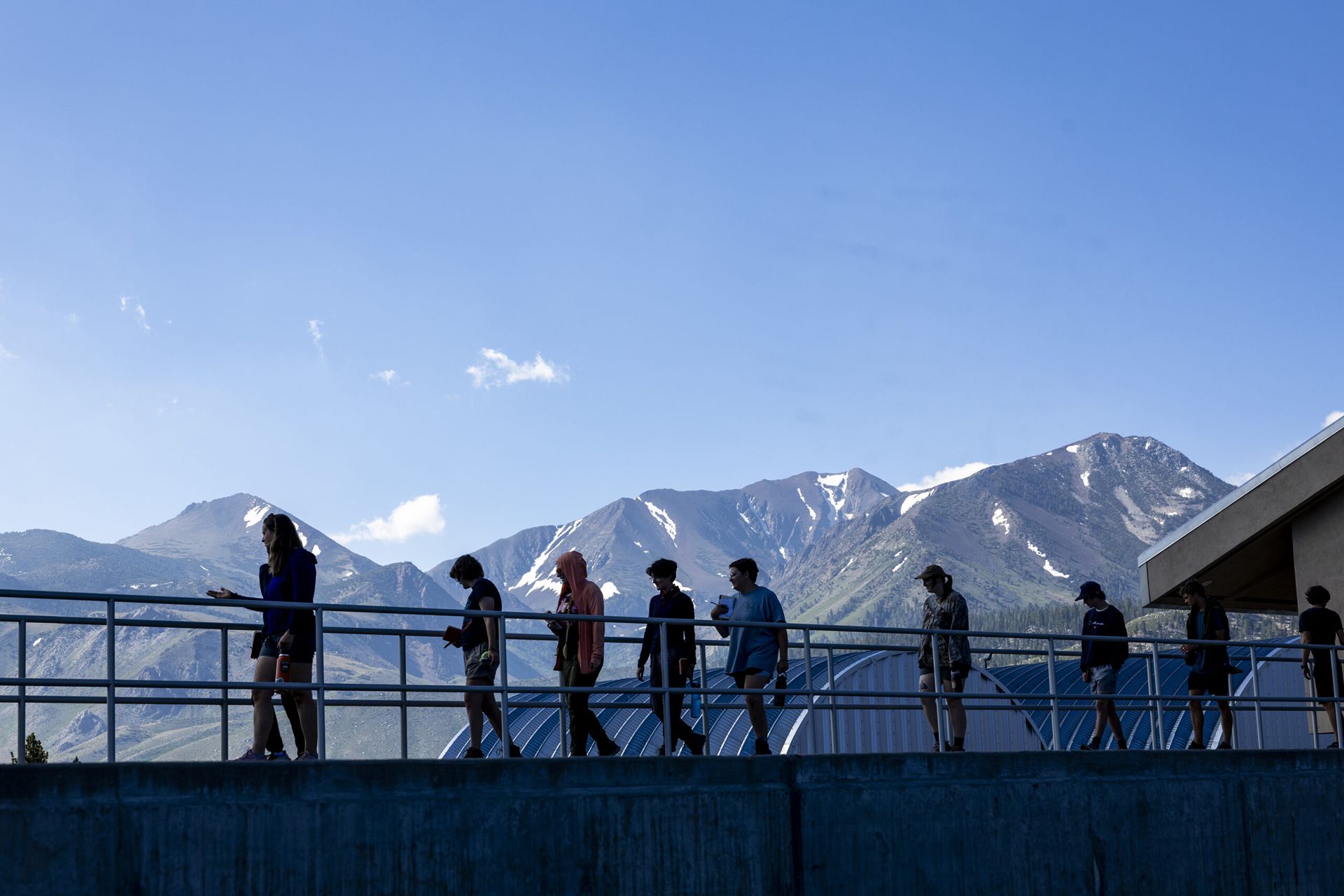 A group of people cross a metal walkway with snow capped mountains in the background.