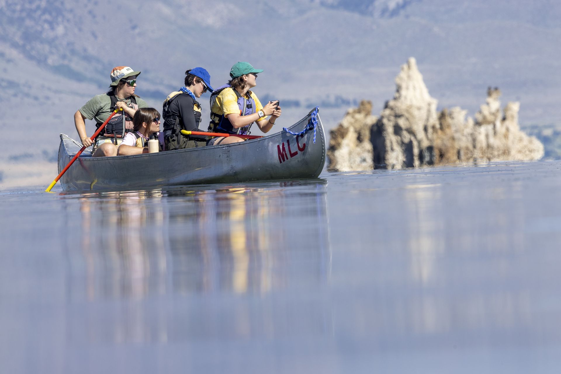 One student paddles s other sits in a canoe on a lake.