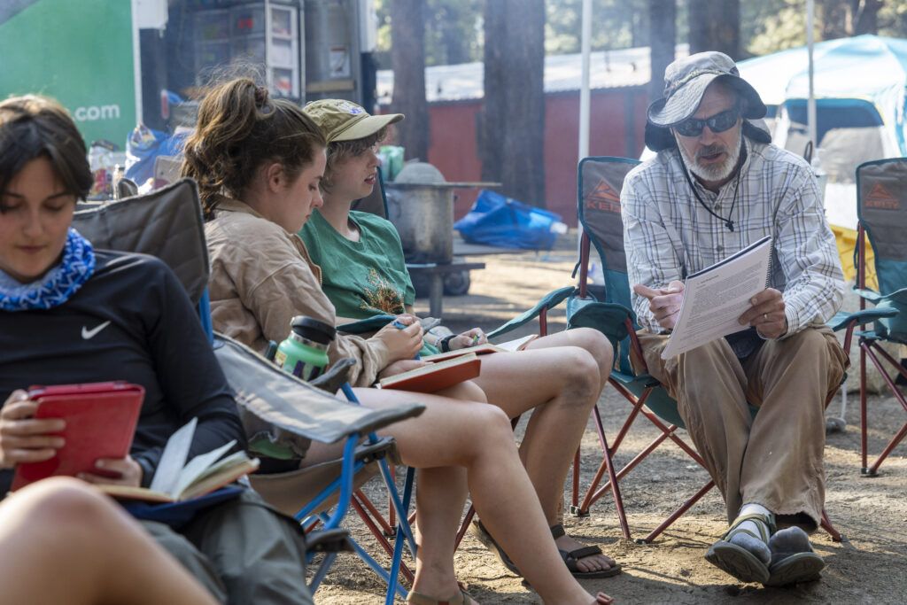 Two students and a professor sit on camp chairs outside working on an assignment.