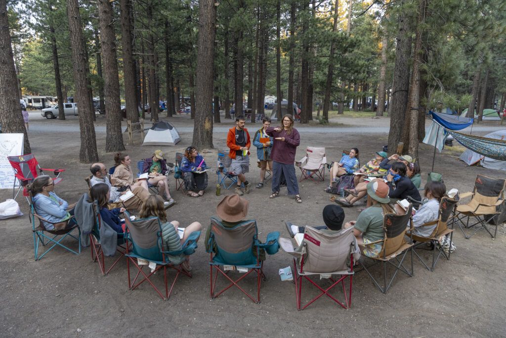 Three students stand at a campground surrounded by  a dozen or so people in camp chairs.