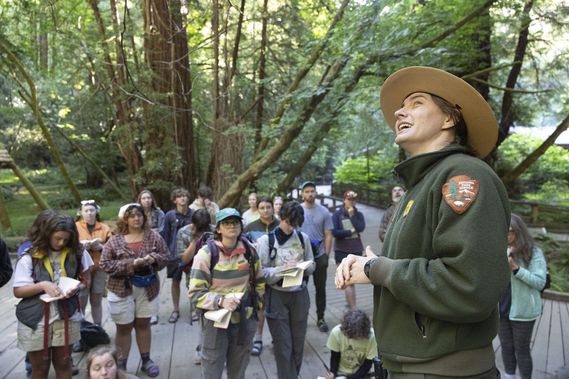 A national park ranger looks up as a class of students take notes in a forest setting.