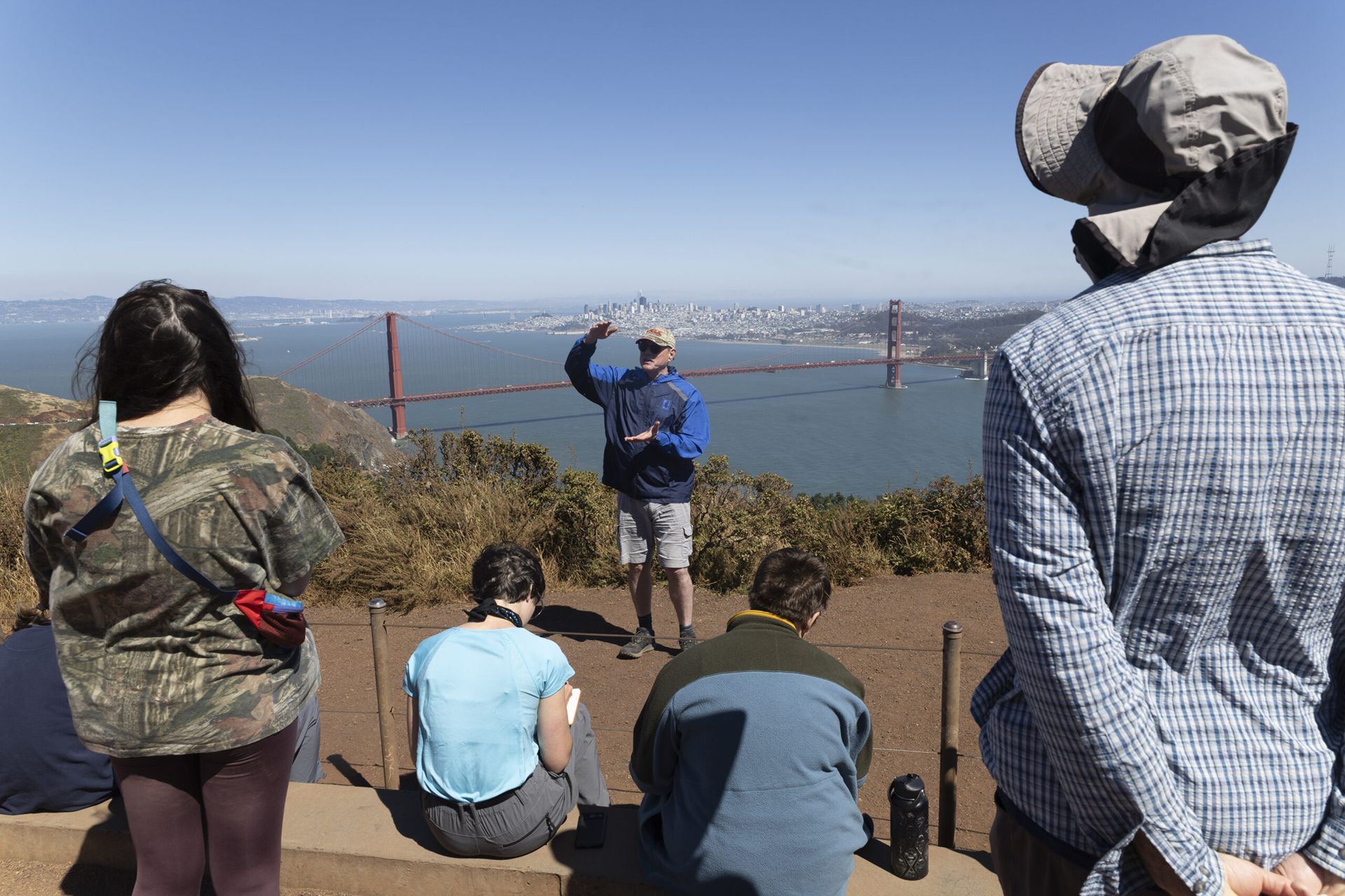 Others look on and take notes as a man in a baseball cap and windbreaker gestures with his hands. The Golden Gate Bridge and San Fransisco's cityscape is in the background.