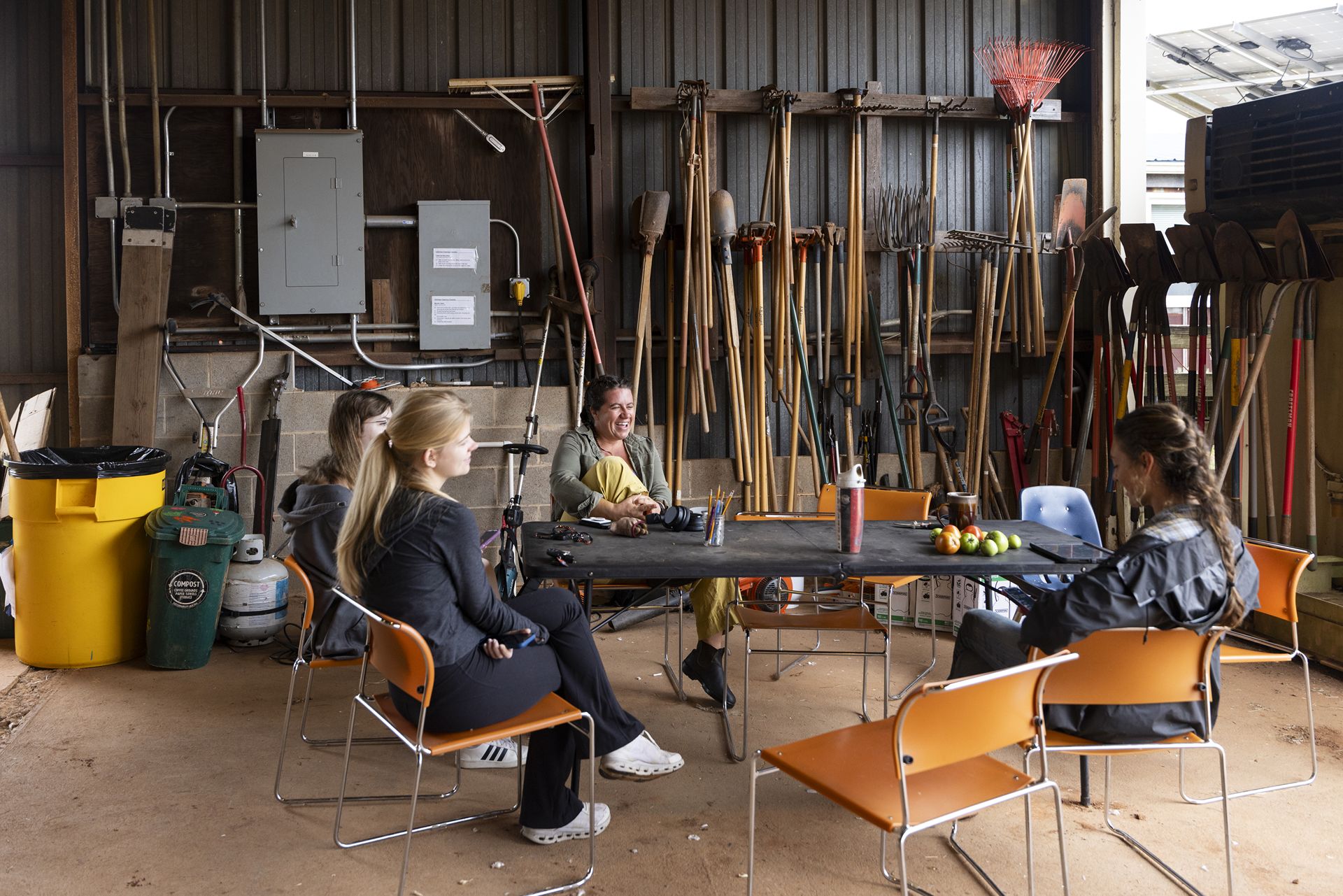 Four women sit at a table in a workshop. The walls are covered in garden tools, including rakes, shovels, and pitchforks.