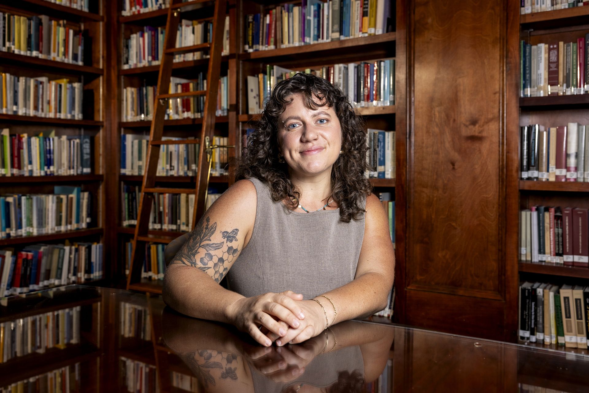 A portrait photo of a young woman in a library. She has curly hair, a bumblebee tattoo on her arm, and is wearing a gray sleeveless top.