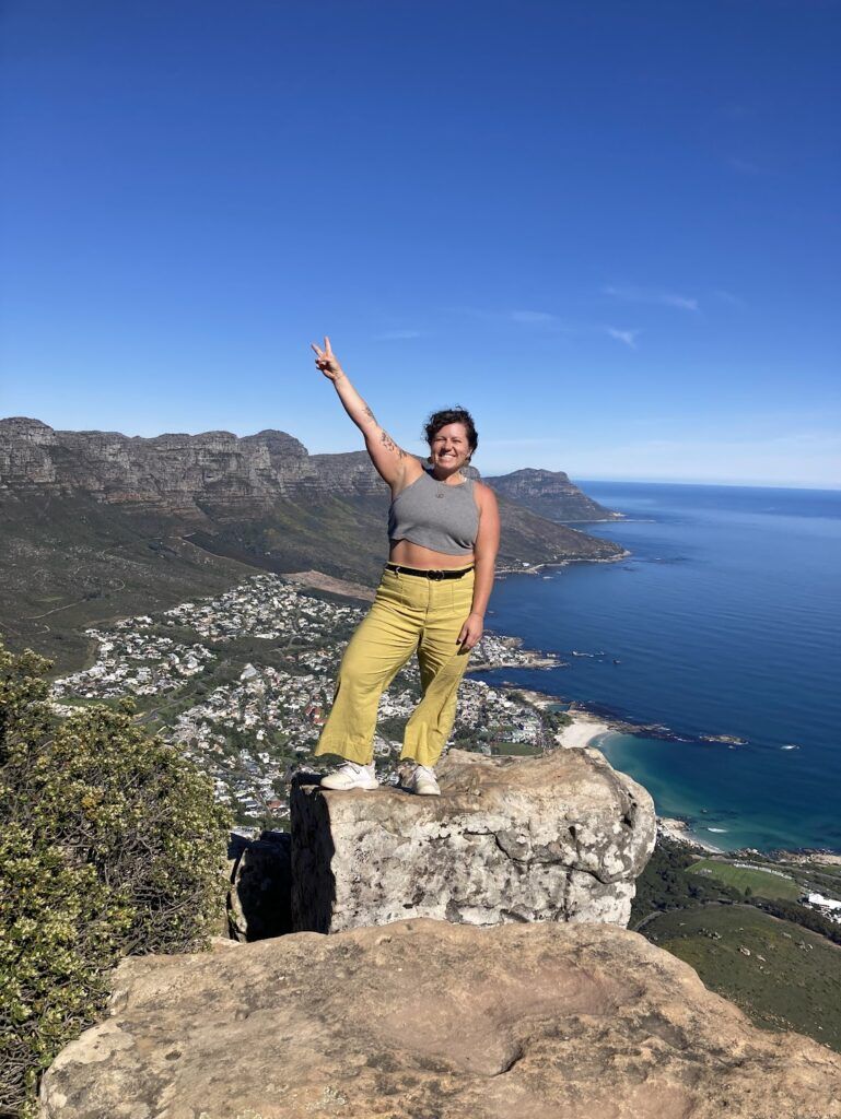 UGA student Julianne O’Connell stands bravely on a small area of rock high above the ocean. She is holding up a peace sign and there is a sunny coastline in the background.