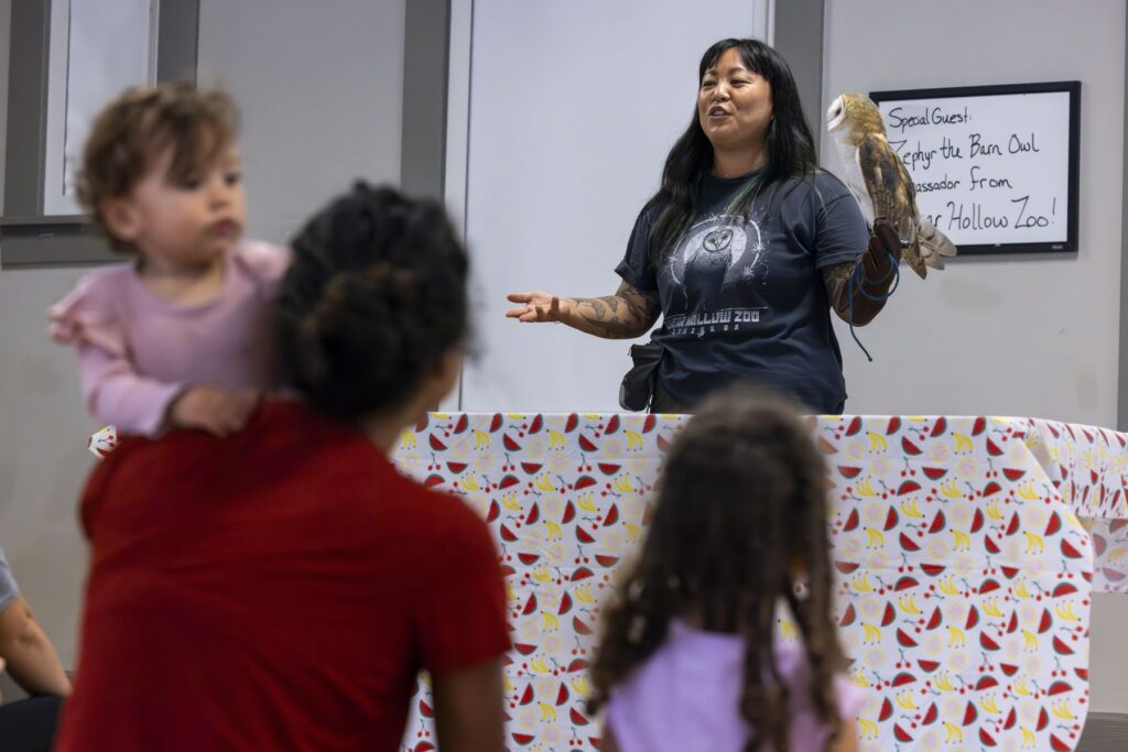 A woman in a grey t-shirt that reads "bear hollow zoo" holds a golden and white owl on her arm as she teaches children at the zoo.