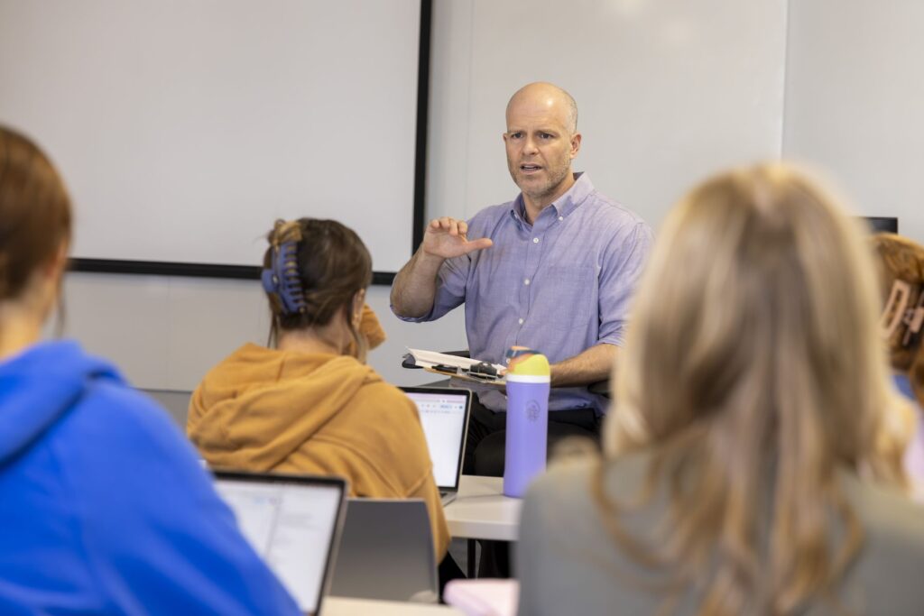 A professor with a serious expression stands at the front of a classroom and teaches a college class. The students can be seen from the back and seem to be intently listening to the lecture.