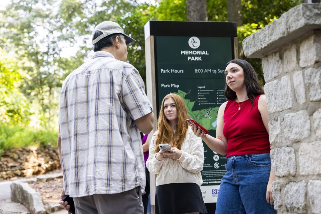 Two students in casual clothing have their phones out as they interview an older man in casual clothing. They are standing in front of a sign that reads "memorial park".