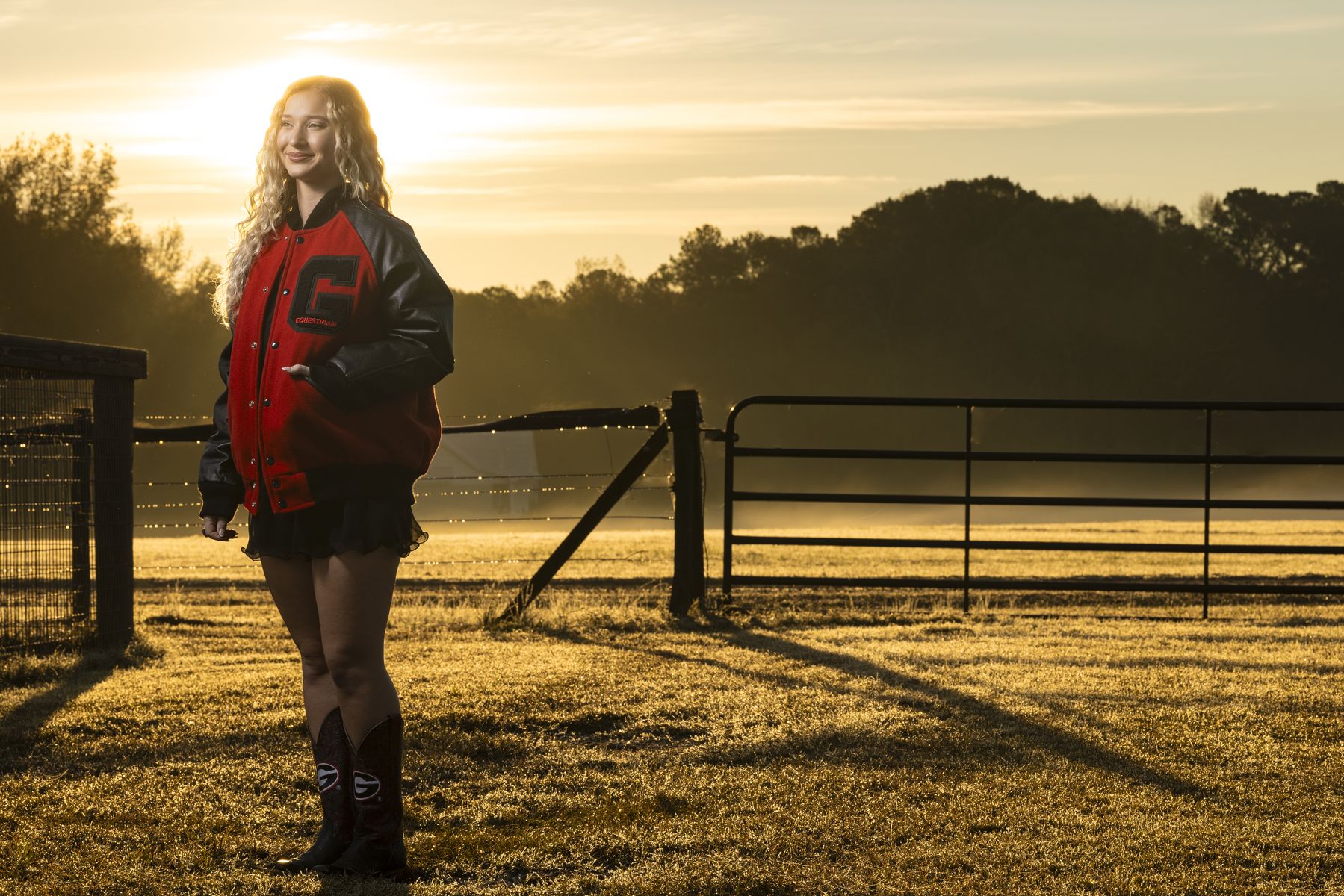 A young woman with blond hair and wearing a black and red University of Georgia letter jacket stands in a large field and looks off into the distance. In the background is long metal gate, a dark forest, and a setting sun.