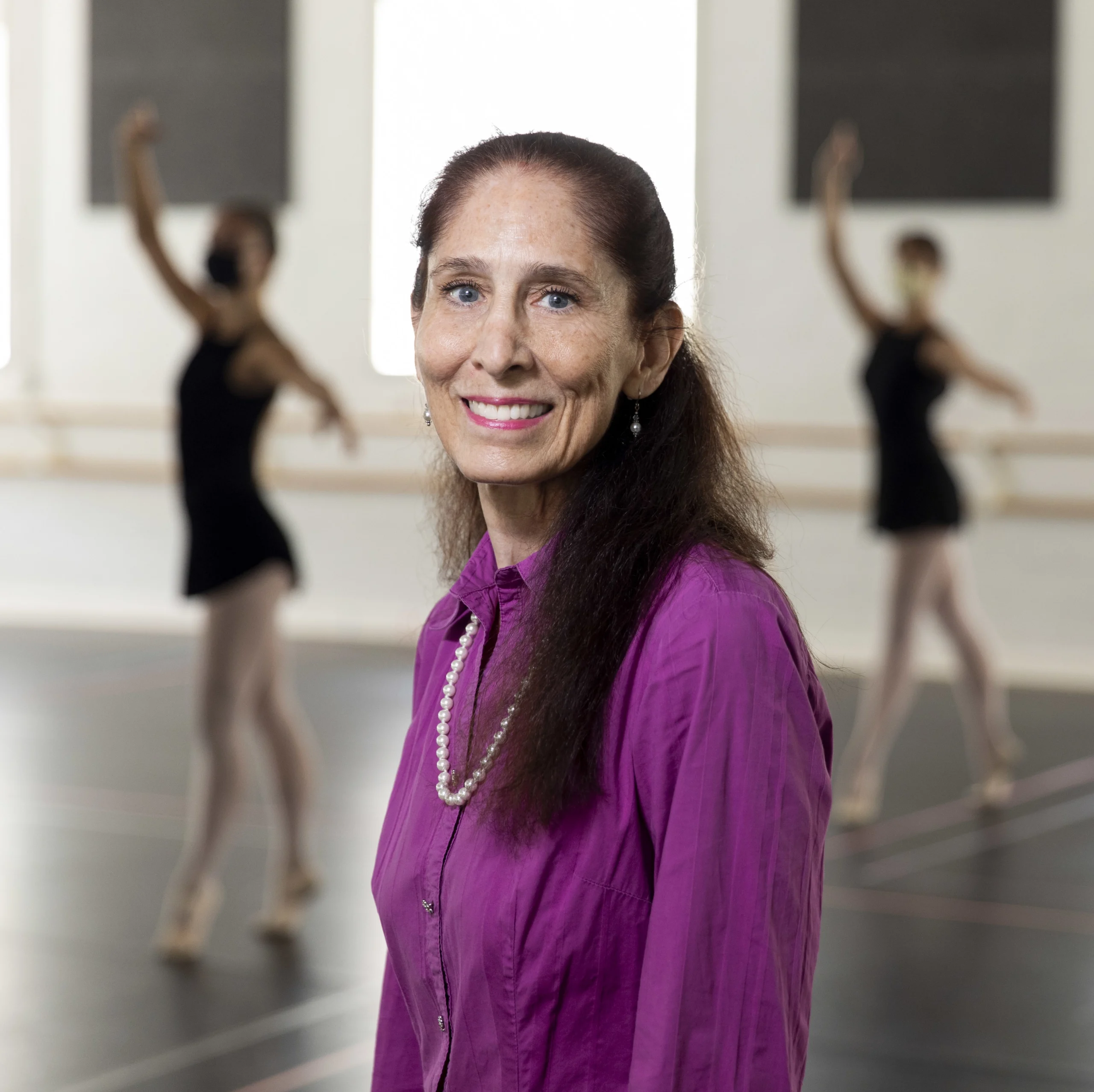 Dance instructor Lisa Fusillo poses in front of two ballet dancers who are blurred in the background. Fusillo, who is smiling, has long dark hair pulled back and wears a magenta blouse and string of pearls. 